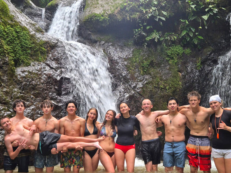 A group of people are standing in front of a waterfall. Most of them are shirtless and wearing swimwear. Some are holding one person horizontally. The background features lush greenery and rocks, with the waterfall cascading down. It appears to be a tropical or subtropical location.